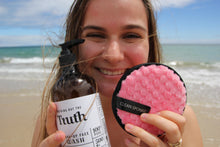 Load image into Gallery viewer, Smiling woman at beach holding Truth Cosmetics java and mandarin face wash bottle and pink cleansing sponge
