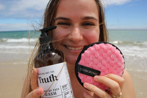 Smiling woman at beach holding Truth Cosmetics java and mandarin face wash bottle and pink cleansing sponge